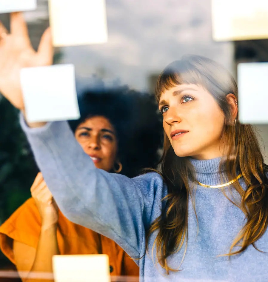 Two women examining colorful sticky notes attached to a glass window in a collaborative workspace.
