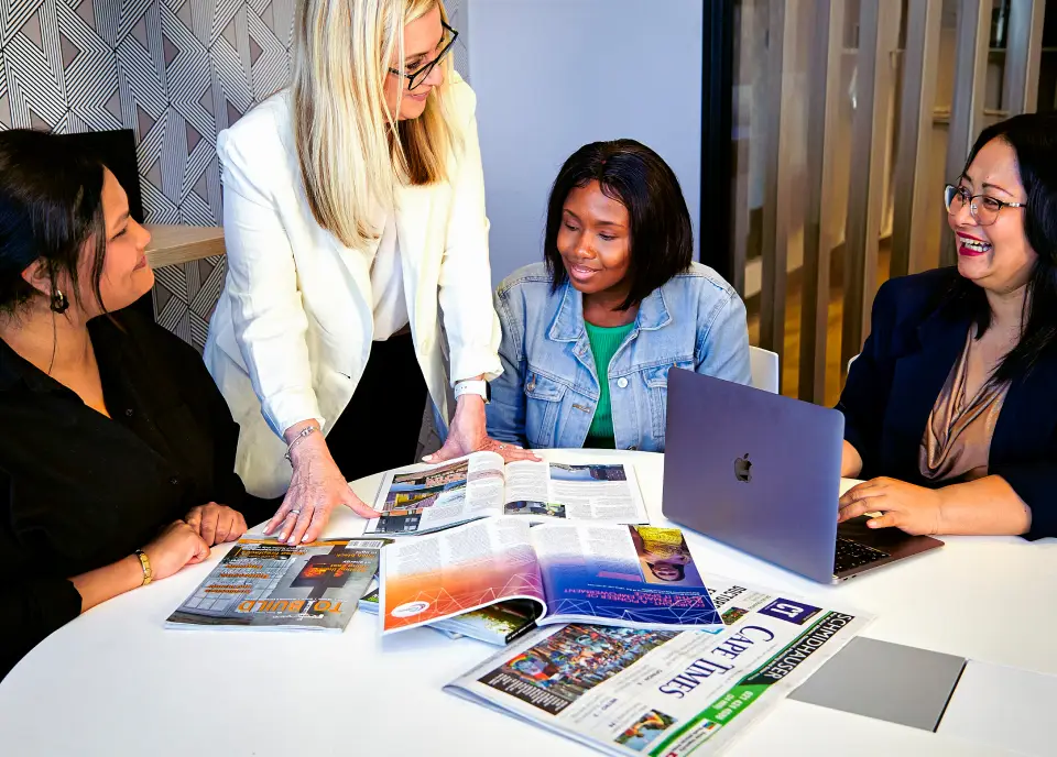 A diverse group of women seated at a table in an office, examining a laptop screen and sharing ideas.